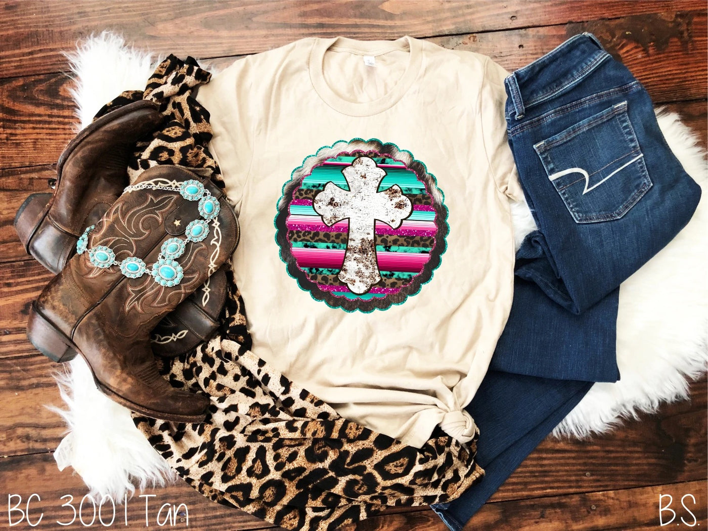 Super soft graphic tee featuring a chippy white cross on a leopard print, pink and teal serape pattern background with glitter effect, and a scalloped cowhide outline. The perfect country western t-shirt to show your faith. 